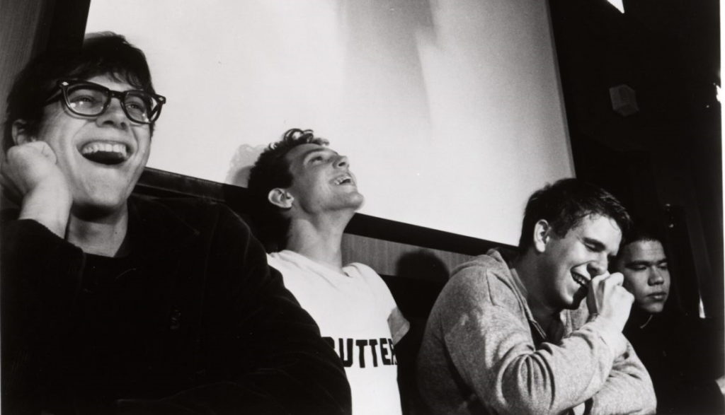 Slint in either 1989 or 1990 sitting and laughing in front of a projector screen. Left to right: Britt Walford, Brian McMahan, Todd Brashear, David Pajo